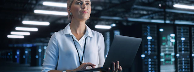 Eine Frau steht mit einem Laptop in einem Serverraum.