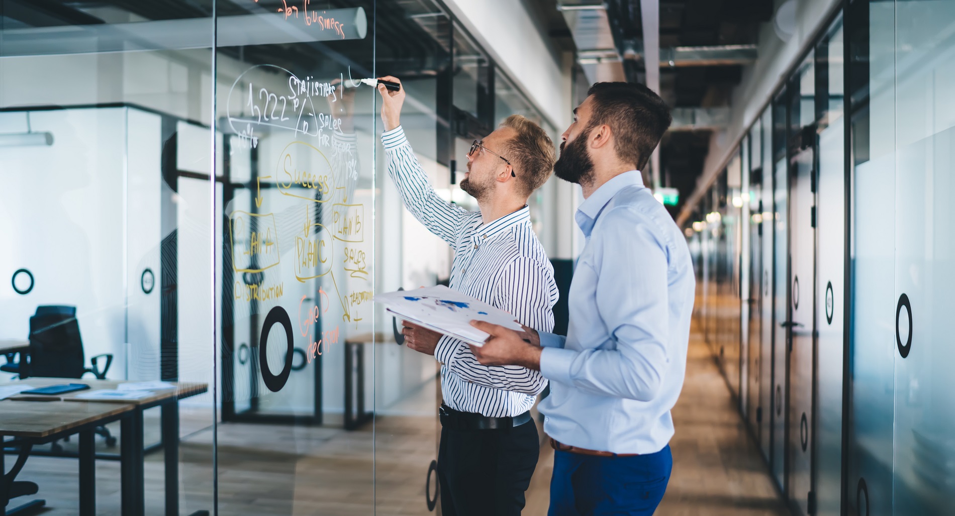 Zwei Männer stehen vor einem durchsichtigen Whiteboard und schreiben darauf