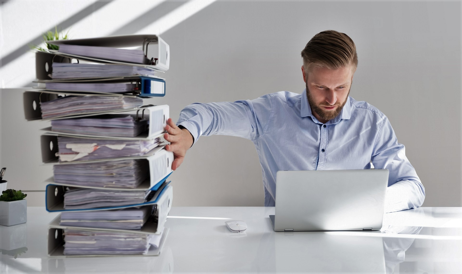 Ein Mann sitzt an einem Schreibtisch vor einem Laptop und schiebt einen Aktenberg beiseite