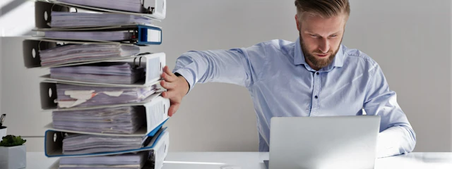 Ein Mann sitzt an einem Schreibtisch vor einem Laptop und schiebt einen Aktenberg beiseite