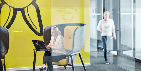 Eine Frau sitzt auf einem Sessel vor einer gelben Wand an ihrem Laptop und daneben läuft eine Person eine Gang entlang