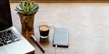Auf einem Tisch liegen ein Notizblock, ein Kugelschreiber und ein Handy, daneben steht eine Tasse Kaffee
